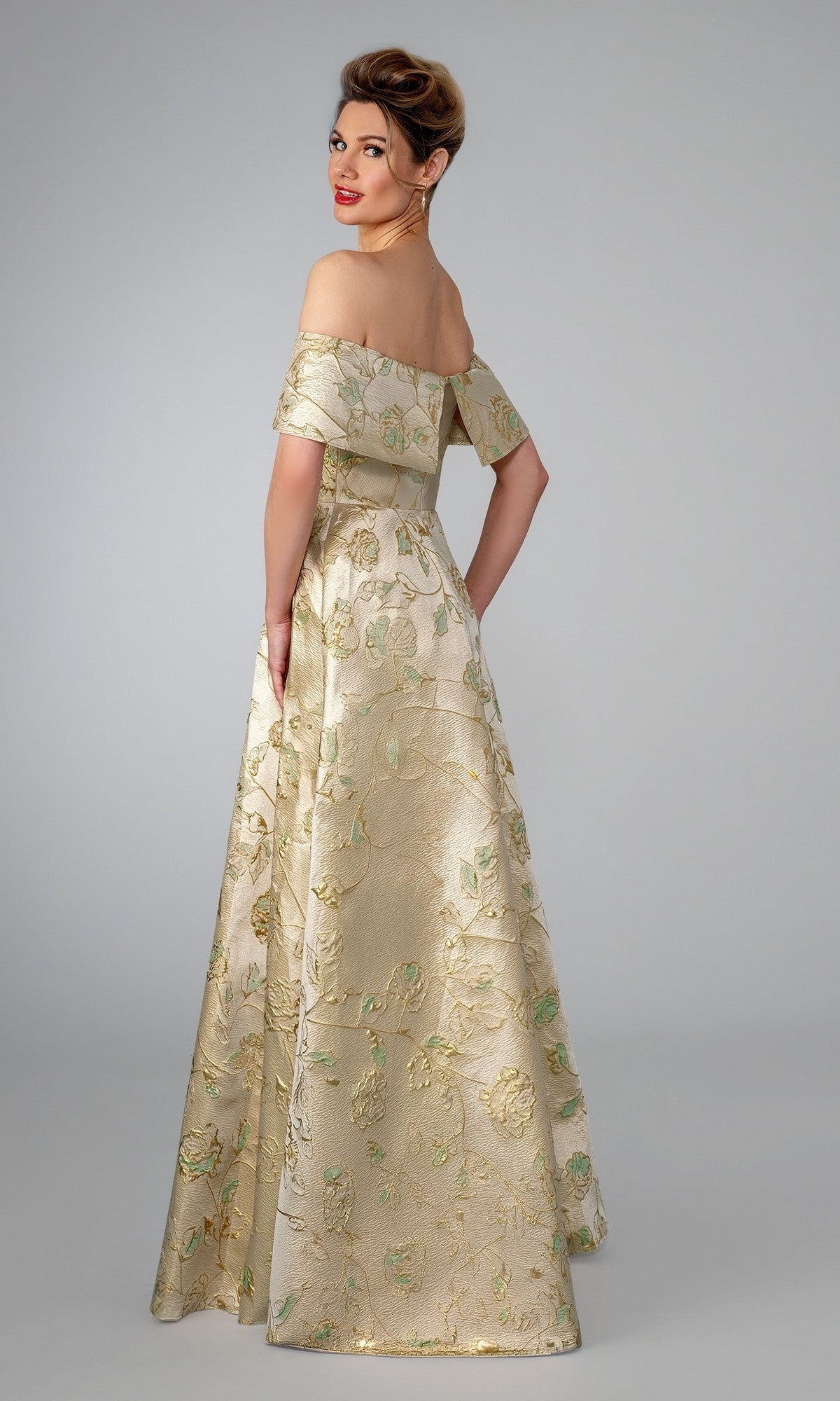 A woman stands facing away, looking back over her shoulder, wearing the Stella Couture 26313 long formal off-shoulder gold brocade gown with floral patterns against a plain background.